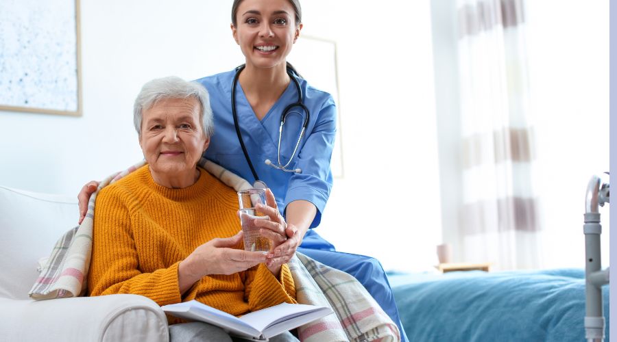Caregiver smiling while assisting an elderly woman holding a glass of water, symbolizing compassion and support as part of a Positive Behaviour Support Plan.